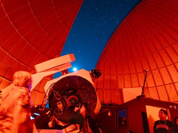 lisa garcia looking through a telescope under a dome lit with red light slightly open to show a starry sky